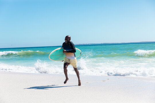 African american carrying surfboard running at beach against blue sky with copy space on sunny day - Powered by Adobe