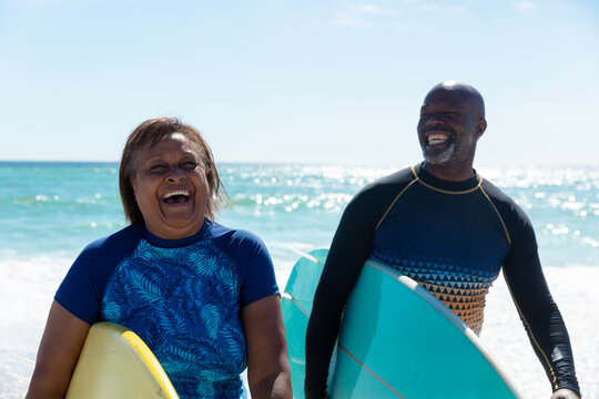 Cheerful african american senior retired couple carrying surfboards enjoying sunny day at beach