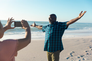 Retired african american senior woman photographing man with arms outstretched at beach - Powered by Adobe