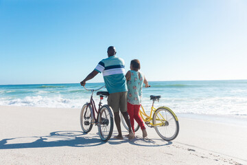 Fototapeta na wymiar Rear view of african american senior couple wheeling bicycles at beach with copy space on blue sky