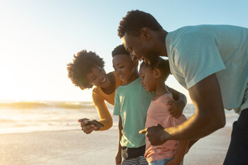 Happy african american woman sharing smartphone with family at beach during sunset
