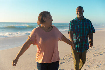 Smiling african american senior woman holding hand of man walking at beach