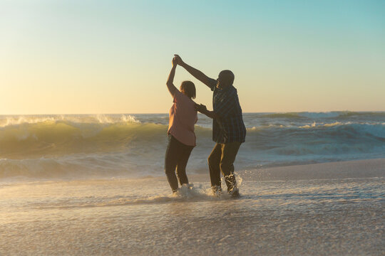 Happy african american retired senior couple dancing on shore at beach against sky during sunset