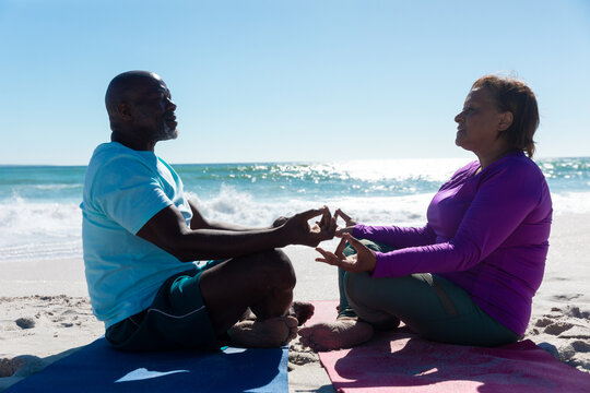 African american senior couple meditating while practicing yoga at beach on sunny day