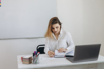 Portrait of a young teacher girl in her classroom. The girl conducts online learning on her laptop with students. Looks in the book Webinar. Online learning