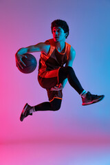 Young energetic man playing basketball isolated on gradient pink blue studio background in neon...