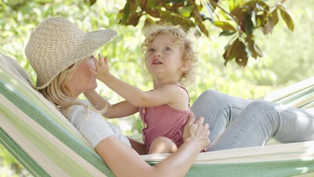 happy mothers day, smiling mom playing with her blue eyed little girl daughter child with curly blonde hair, lying on hammock  little girl kisses mom in sunny garden, spring and summer time concept