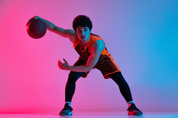 Young energetic man playing basketball isolated on gradient pink blue studio background in neon light. Youth, hobby, motion, activity, sport concepts.