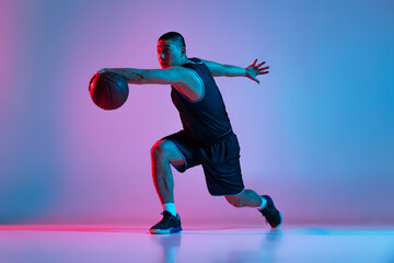Young sportive man, basketball player training with ball isolated on blue studio background in neon light. Youth, hobby, motion, activity, sport concepts.