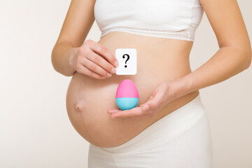 Young adult pregnant woman hands holding and showing pink blue egg and white card of question mark on big naked belly. Guessing future baby gender. Baby expectation. Isolated on light gray background.
