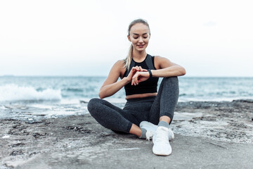 Tired but smiling fit woman in sportswear sitting on urban beach and look at smart bracelet