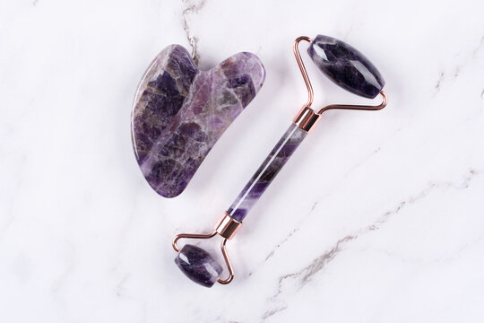 Facial massage kit - amethyst gua sha tool and face roller on white marble background. Face roller, massager made from natural stones, home spa concept