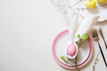 table setting for Easter. pastel dusty pink egg in napkin folded in shape of easter bunny on plate decorated with live blossoming branch with leaves. background with copy space