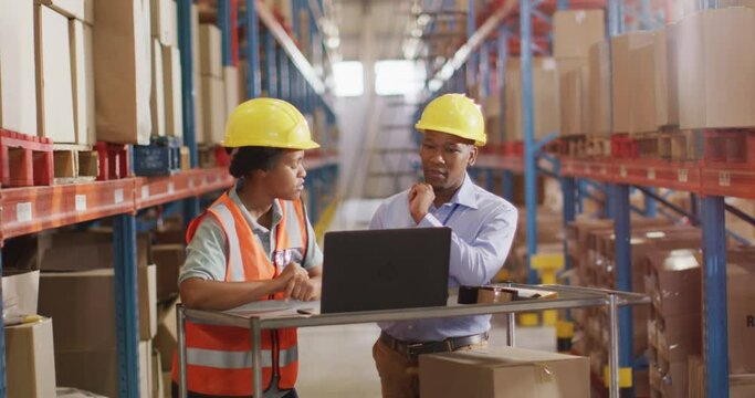 African american male and female workers wearing helmet and using laptop in warehouse