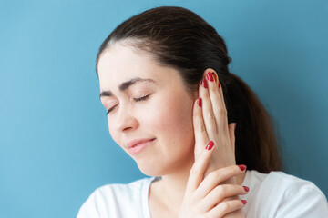 Portrait of a disgruntled young Caucasian woman covering her ear from pain or a loud sound. Blue background. The concept of deafness and ear diseases