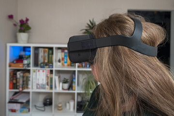 Fototapeta premium Close-up of a girl's head wearing VR glasses against a rack of games and flowers. Side view. Woman traveling in the metaclasses in her home. A virtual reality helmet
