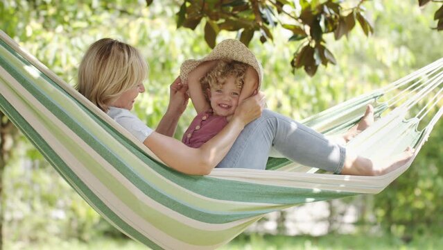 happy mothers day, smiling mom playing with her blue eyed little girl daughter child, plays with her by putting on a big straw hat on hammock in sunny green garden, spring and summer time concept
