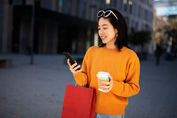 Pretty chinese girl using mobile phone and holding coffee to go. Beautiful woman enjog in coffee while walking on the city street.