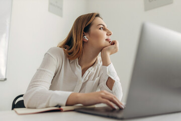 A bored young woman sits at her home office desk, looks at a distance, cannot work at a laptop, an exhausted student feels unmotivated, unwilling to learn, distracted by a break. 