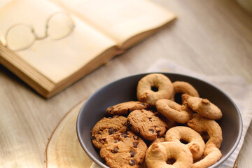 Plate of chocolate chip cookies and sugar cookies, vintage books and reading glasses, candle and flower on the table. Hygge at home. Selective focus.