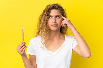 Young caucasian woman holding a brushing teeth isolated on yellow background having doubts and thinking