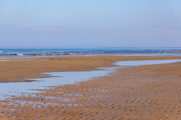 Low angle view to sandy beach partly covered by water, in distance wavy sea and light blue sky. Seashore at Majori, Jurmala, Latvia at early spring day. Empty paradise beach in north.