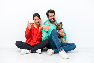 Young caucasian couple sitting on the floor with their pet isolated on white background making rock gesture