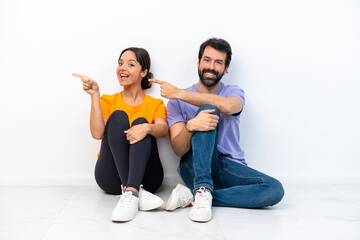 Fototapeta na wymiar Young caucasian couple sitting on the floor isolated on white background pointing finger to the side and presenting a product