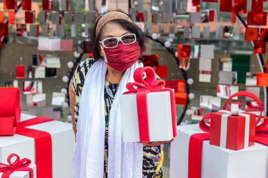 Attractive Senior Woman With Gift In Hand. Portrait An Older Woman With A Gift Box. Elderly Woman Wear Glasses And Fabric Mask Holding A Gift Box