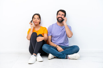 Fototapeta na wymiar Young caucasian couple sitting on the floor isolated on white background talking to mobile