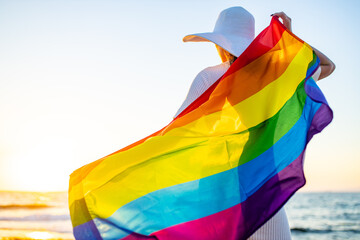 woman with straw white hat holding a gay pride rainbow flag blowing in the wind on beach with golden sunset