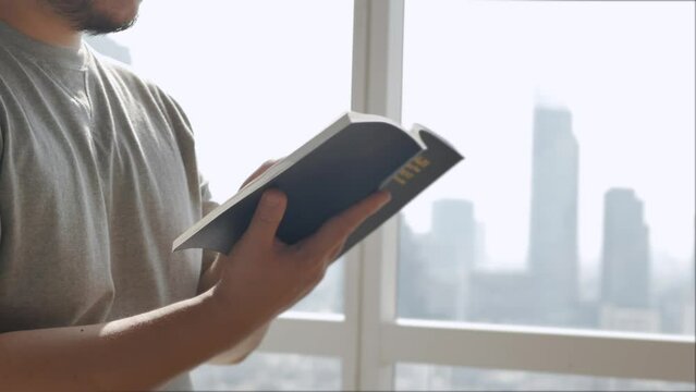an asian man reading a booking in living room with background of the city center with skyscraper through big window