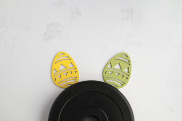 Dumbbells barbell weight plate disc and decorative Easter eggs in shape of bunny ears. Healthy fitness lifestyle rabbit composition, gym workout and training concept. Fit flat lay with copy space.