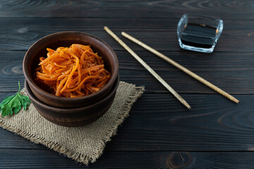 Korean carrot salad in a clay salad bowl on a canvas napkin. Wooden chopsticks on the table. Soy...