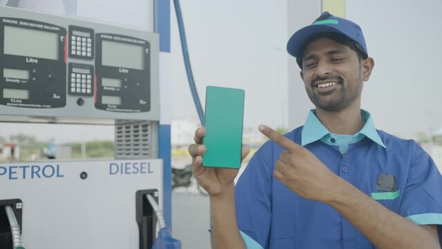 Smiling petrol pump worker showing green screen mobile phone by pointing finger by looking at camera at filling station - concept of online booking, advertisement and gasoline servie.