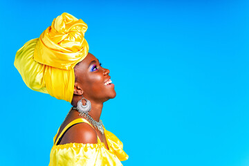 beautiful african woman in yellow silk turban on head and dress with silver neckless and earrings , nose piercing in blue studio background - obrazy, fototapety, plakaty