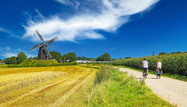 View on cycling path with cyclists couple in rural dutch limburg maas landscape with windmill (Molen de grauwe beer) against blue summer sky - Beesel, Netherlands