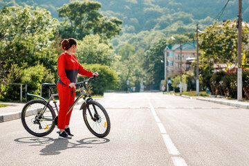 Sports and activity. Full-length portrait of a beautiful young woman in red sportswear, crossing the road on a Bicycle. Side view. In the background, a street and a road in the sunlight. Copy space