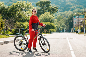 Sports and activity. Full-length portrait of a smiling young beautiful woman in red sportswear, crossing the road on a Bicycle. In the background, a street and a road in the sunlight