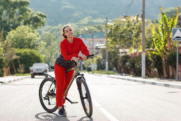 A young beautiful smiling woman in sportswear stands next to a Bicycle. In the background, the empty road and mountains, a Sunny day. Sports lifestyle and Cycling. Copy space