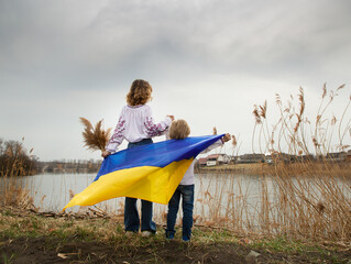 little boy and a woman stand with their backs on the shore of lake with blue and yellow Ukrainian...
