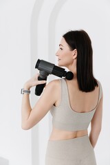 A girl in a beige sports suit massaging her back. vibrating massager close-up. Electric massager pistol in hand massaging the muscles. Sports recovery concept. 