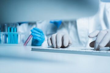 partial view of bioengineer in latex gloves typing on computer keyboard near colleague with test tube.