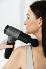 A girl at home massaging with a vibrating massager with a nozzle.  An electric therapeutic pistol massager in her hand massages the muscles of her arm. Sports recovery concept after a workout. 
