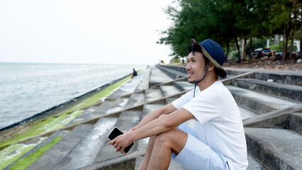 Good looking Asian guy, sitting alone on a beautiful beach, enjoying the cool breeze, popular tourist destination