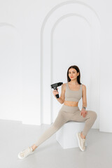 A girl in a tracksuit is holding a vibrating massager.  An electric pistol massager in her hand massaging the muscles. Sports recovery concept after a workout. 