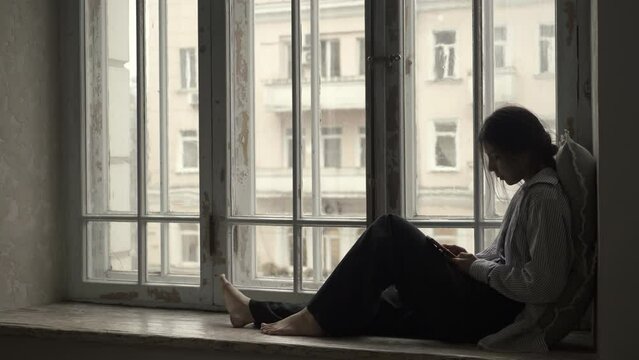 Concentrated beautiful asian young woman sitting on window sill and browsing social media copy space. Pretty woman communicates remotely, text messages on smartphone. Filmed in cinematic style