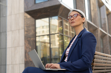 Pensive mature businesswoman wearing stylish eyeglasses using laptop computer working online on the street, copy space. Confident asian freelancer sitting at workplace looking away