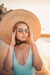 A young tanned woman in a beautiful swimsuit with a straw hat stands and rests on a tropical beach with sand and looks at the sunset and the sea. Selective focus. Vacation concept by the sea