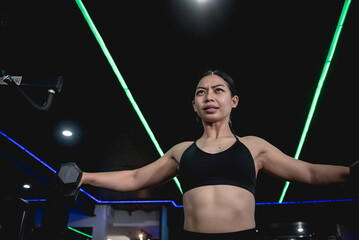 A fit asian woman grimaces while feeling the lactic acid burn during a high rep dumbbell lateral...